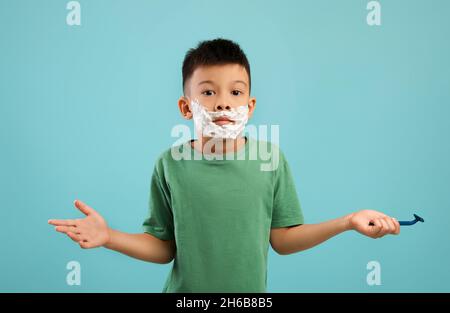 Portrait de drôle petit garçon asiatique avec mousse de rasage sur le visage haussant les épaules dans la confusion, mignon Korean Kid tenant Razor et regardant le Camer Banque D'Images