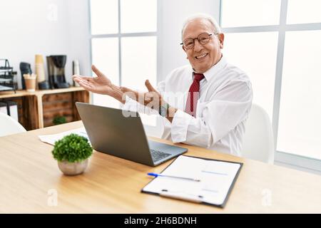 Homme senior travaillant au bureau à l'aide d'un ordinateur portable invitant à entrer dans le sourire naturel avec la main ouverte Banque D'Images