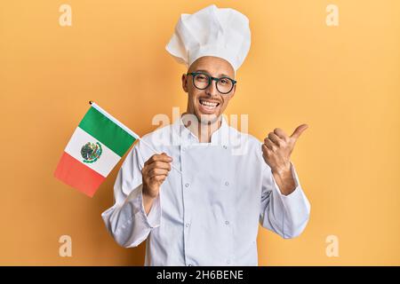 Homme chauve avec barbe portant un tablier de cuisinier professionnel tenant le drapeau du mexique pointant le pouce vers le haut du côté souriant heureux avec la bouche ouverte Banque D'Images