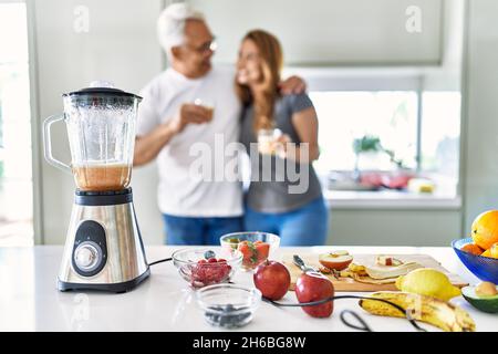Couple hispanique d'âge moyen souriant heureux et embrassant boire un verre de smoothie à la cuisine. Banque D'Images