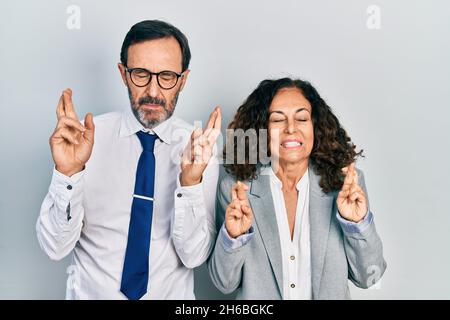 Un couple d'âge moyen de femme et d'homme hispaniques portant un uniforme de bureau d'affaires gesturant le doigt croisé souriant avec espoir et les yeux fermés. Chance et super Banque D'Images