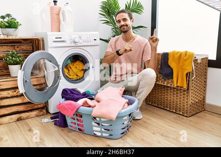 Jeune hispanique mettant le linge sale dans le lave-linge souriant et regardant la caméra pointant avec deux mains et les doigts sur le côté. Banque D'Images