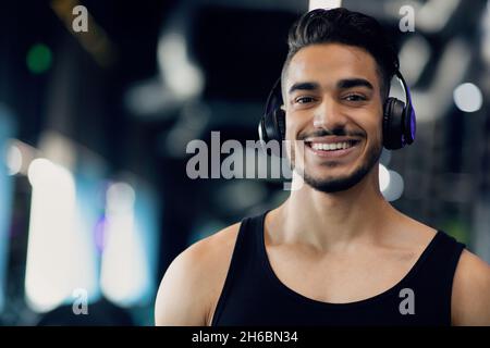 Portrait de Happy Athletic Arab Man portant des écouteurs sans fil posant dans la salle de gym, beau Moyen-Orient Guy écoutant de la musique pendant l'entraînement dans le jeu de sport Banque D'Images