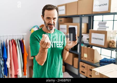 Homme d'âge moyen avec la barbe portant t-shirt volontaire tenant smartphone faisant le geste d'argent avec les mains, demandant le paiement de salaire, millionnaire affaires Banque D'Images