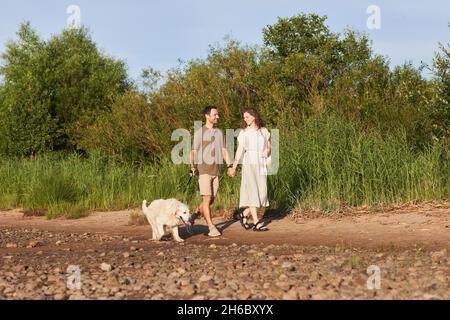 Portrait grand angle de couple heureux avec chien marchant ensemble par l'eau dans la lumière du soleil, espace de copie Banque D'Images