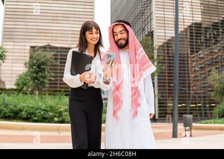 Faible angle de femme d'affaires gaie dans un homme arabe à keffiyeh souriant et utilisant un smartphone ensemble tout en ayant des réunions d'affaires dans la rue de la ville moderne Banque D'Images