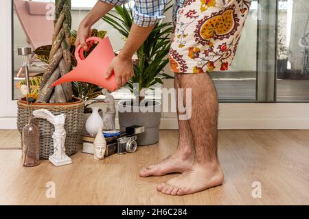 Homme portant des shorts arrosoir plantes avec un arrosoir dans un salon avec des figures décoratives, caméra, vase à côté d'une grande fenêtre menant à un terr Banque D'Images