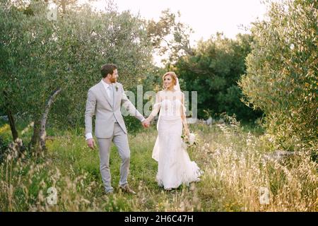 La mariée et le marié marchent en tenant les mains parmi les arbres dans le parc Banque D'Images