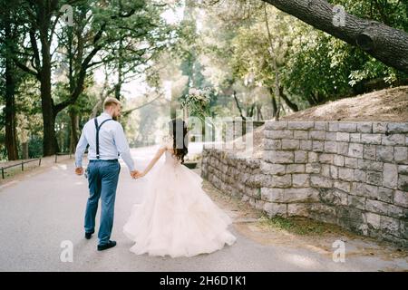 La mariée et marche en tenant les mains sur la route près du mur de pierre entre les arbres dans une oliveraie, vue arrière Banque D'Images