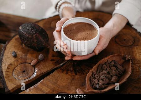 Cacao de cérémonie fait main chaud dans une tasse blanche.Femme mains tenant cacao d'artisanat, vue de dessus sur table en bois.Boisson au chocolat biologique saine préparée à partir de Banque D'Images