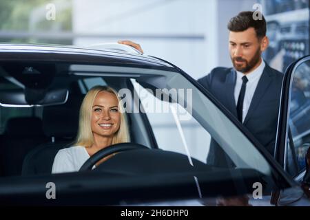 Vendeur compétent en costume présentant une nouvelle voiture de luxe pour femme assise à l'intérieur.Une cliente caucasienne consulte le concessionnaire tout en achetant une nouvelle voiture dans le hall d'exposition. Banque D'Images