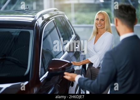 Belle blonde souriante se tenant près d'une voiture de luxe tout en parlant avec un vendeur au salon.Manager compétent pour aider les femmes à choisir un nouveau véhicule. Banque D'Images