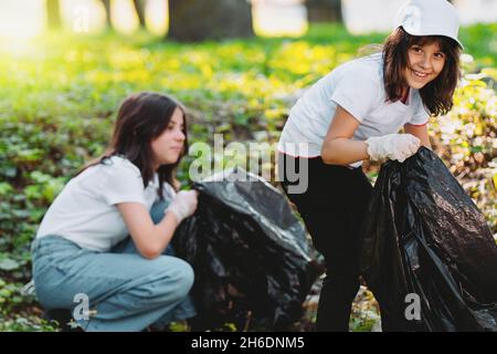 Jolie fille volontaire regardant l'appareil photo rassembler des ordures en utilisant le sac poubelle portant des gants de protection et une autre fille faisant le même travail Banque D'Images