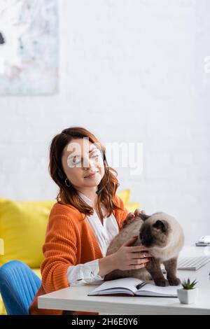femme souriante dans un micro-casque travaillant à la maison et chat de petting assis sur le bureau Banque D'Images