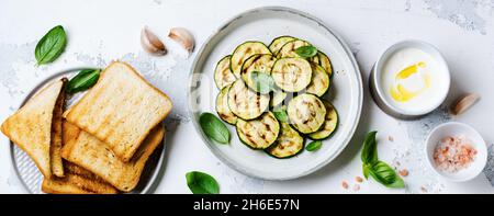 Salade de courgettes grillées aux feuilles de basilic, sauce au yaourt et pain frit dans une simple plaque en céramique sur fond de béton blanc.Flat lay avec copie sp Banque D'Images