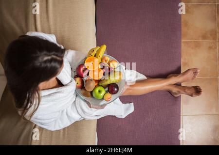 Photo ci-dessus de femme en peignoir blanc assis sur le lit avec une assiette de fruits colorés Banque D'Images