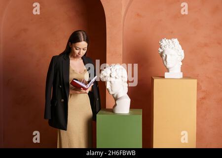 Portrait d'une femme charmante, concentrée et attentive, avec carnet dans ses mains, dans une robe dorée et une veste noire à côté d'un buste en plâtre, planification et analyse, concept publicitaire Banque D'Images