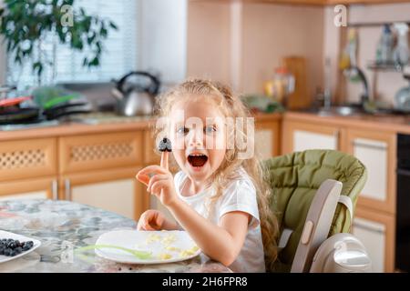 La petite fille mange du porridge avec des baies dans la cuisine et joue avec des baies les mettant sur votre doigt.Enfant s'amuser et manger le déjeuner à la maison à l'intérieur Banque D'Images