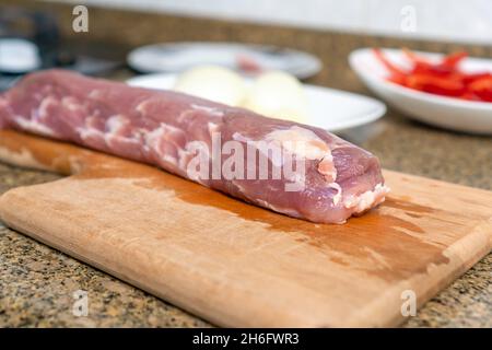 Un morceau de viande de porc sans os repose sur une planche à découper, en arrière-plan d'une assiette avec d'autres ingrédients Banque D'Images
