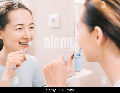 Une jeune femme avec des bretelles nettoie ses dents avec une brosse à dents, avant le miroir.Mise au point sélective sur le miroir. Banque D'Images