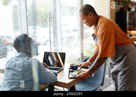 Jeune femme afro-américaine portant un tablier travaillant dans un café apportant un café et un dessert délicieux à un jeune client Banque D'Images