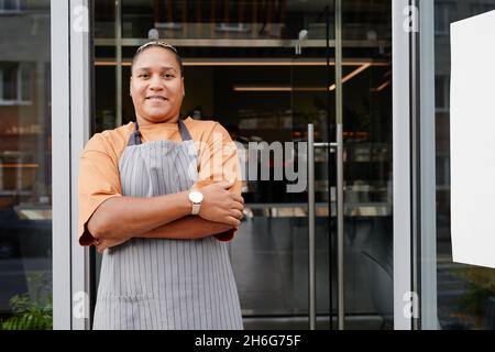 Portrait horizontal moyen de la jeune femme noire portant un tablier debout détendu avec les bras croisés à l'entrée du restaurant souriant à la caméra Banque D'Images
