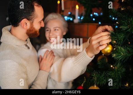 Photo moyenne de jeune couple gai dans l'amour décorant l'arbre de Noël avec des boules brillantes près de la cheminée dans la salle de séjour confortable sombre. Banque D'Images