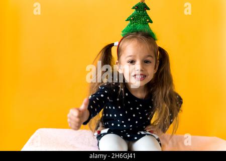 Portrait de sourire positif surpris bonne petite écofille fille Noël arbre décoration sur la tête polka robe à pois montrer pouces vers le haut fond orange Banque D'Images