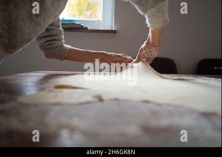 Vue à angle bas d'une femme roulant et tirant une pâte de strudel phyllo maison sur une table à manger maison. Banque D'Images