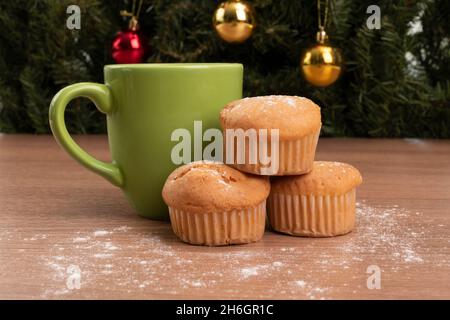 Tasse verte avec boisson à côté de trois cupcakes cuits sur une table en bois décorée de sucre, en arrière-plan un arbre de Noël avec des ampoules colorées Banque D'Images
