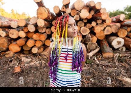 Jeune fille biraciale avec des tresses arc-en-ciel de boîte se tient devant une pile de billes.Elle porte une robe à rayures arc-en-ciel. Banque D'Images