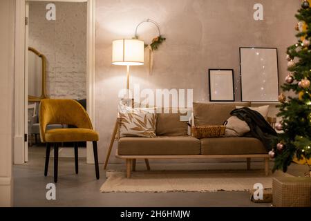 Canapé confortable avec coussins placés sur un tapis près d'un fauteuil dans la salle de séjour avec lampe au sol et arbre de Noël décoré pendant la saison des fêtes Banque D'Images