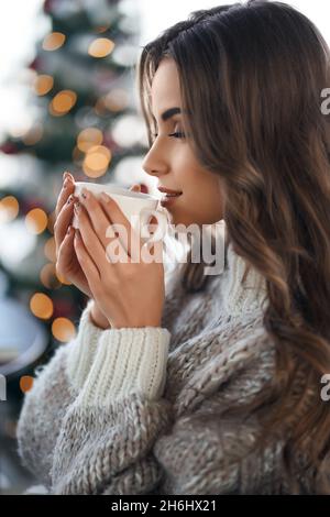 Vue latérale d'une belle femme en pull en dégustant une boisson chaude dans une maison confortable.Arrière-plan flou de l'arbre de noël.Détente pendant les vacances d'hiver. Banque D'Images