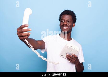 Homme afro-américain qui prétend prendre le selfie sur son vieux téléphone fixe.Personne noire s'amusant avec le téléphone rétro, plaisantant de prendre des photos et souriant, en utilisant le téléphone vintage dans la main Banque D'Images