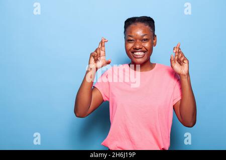 Portrait d'un adolescent afro-américain souriant en croquant les doigts, faisant un souhait optimiste debout en studio sur fond bleu.Jeune femme priant pour Bonne chance.Concept d'attente Banque D'Images