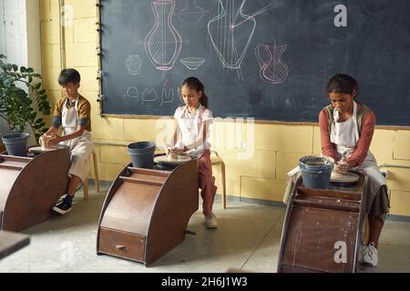 Rangée de trois écoliers interculturels qui font des pots d'argile ou d'autres faïence tout en étant assis à roues à la classe de poterie Banque D'Images