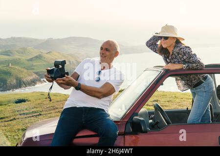 Couple âgé prenant une photo polaride et voyageant à travers la côte dans leur voiture décapotable 4x4 Banque D'Images