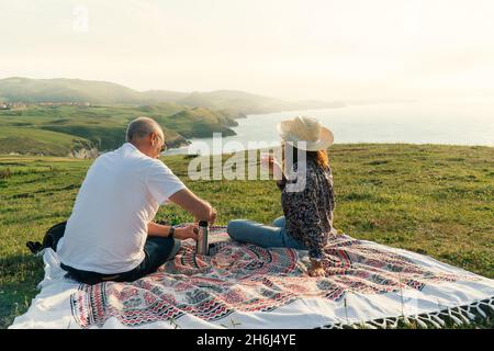 Couple âgé appréciant le coucher du soleil et prenant une tasse de thé chaud sur le rivage Banque D'Images