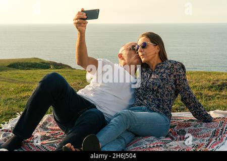 Couple âgé couché sur la prairie, prenant quelques selfies et appréciant le coucher du soleil sur la côte Banque D'Images
