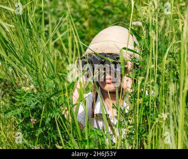 Préadolescence fille portant un casque de liège se cachant dans l'herbe regardant à travers des jumelles.Concept de découverte Banque D'Images