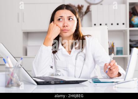 Inquiète femme médecin travaillant à l'ordinateur portable avec des papiers dans le bureau Banque D'Images