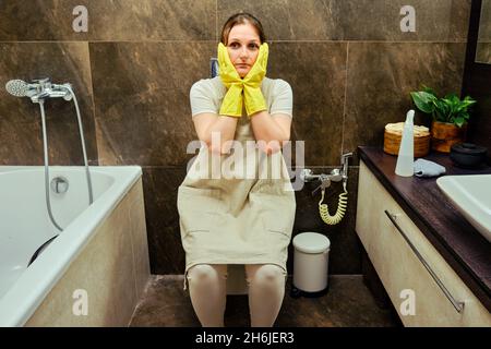 Problème dans une femme triste assise sur les toilettes pendant le nettoyage de la salle de bains.Désinfection avec des gants en caoutchouc jaune dans les toilettes Banque D'Images