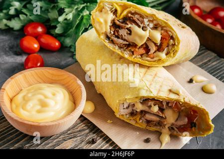 Rouleau de fromage de shawarma avec sauce au fromage, poulet et légumes dans du pain pita sur fond de bois foncé avec tomates cerises et persil vert Banque D'Images