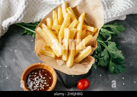 Une grande portion de frites se trouve dans du papier artisanal avec une sauce barbecue, des graines de sésame, des tomates cerises et du persil vert sur une surface en béton foncé avec Banque D'Images