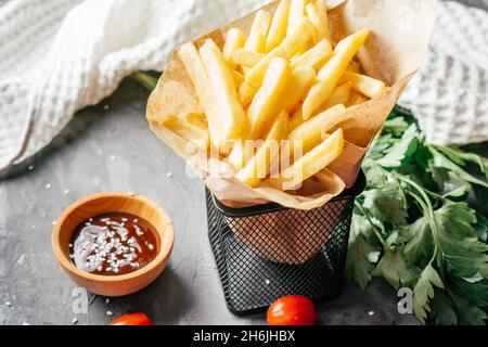 Une grande portion de frites se trouve dans du papier artisanal avec une sauce barbecue, des graines de sésame, des tomates cerises et du persil vert sur une surface en béton foncé avec Banque D'Images