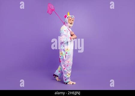 Photo de charmant homme rigolo pensionné unicorn vêtements de nuit marche attrapant papillons souriant isolé couleur violet fond Banque D'Images