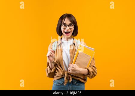 Idée créative.Une étudiante excitée pointe un crayon vers le haut, tenant des cahiers sur fond jaune de studio Banque D'Images