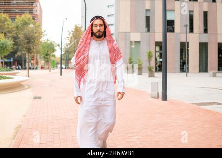 Beau homme arabe réussi sheikh portant des vêtements traditionnels marchant dans la rue de la ville de Dubaï, homme d'affaires arabe confiant dans le blanc saoudien kandora en plein air Banque D'Images