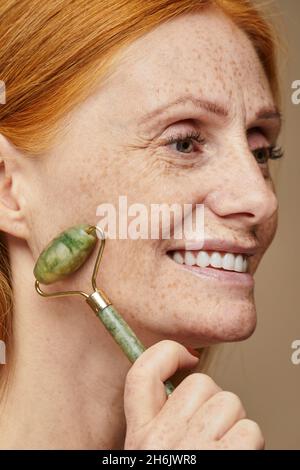 Portrait vertical de belle femme aux cheveux rouges avec des taches de rousseur souriant et faisant un massage du visage à l'aide de rouleau de pierre Banque D'Images