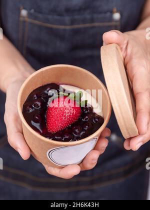 Les mains du chef pâtissier tiennent la tarte aux bleuets et aux fraises fraîches dans une tasse en papier avec étiquette vide.Gâteau au fromage myrtille fait maison dans une tasse en papier. Banque D'Images